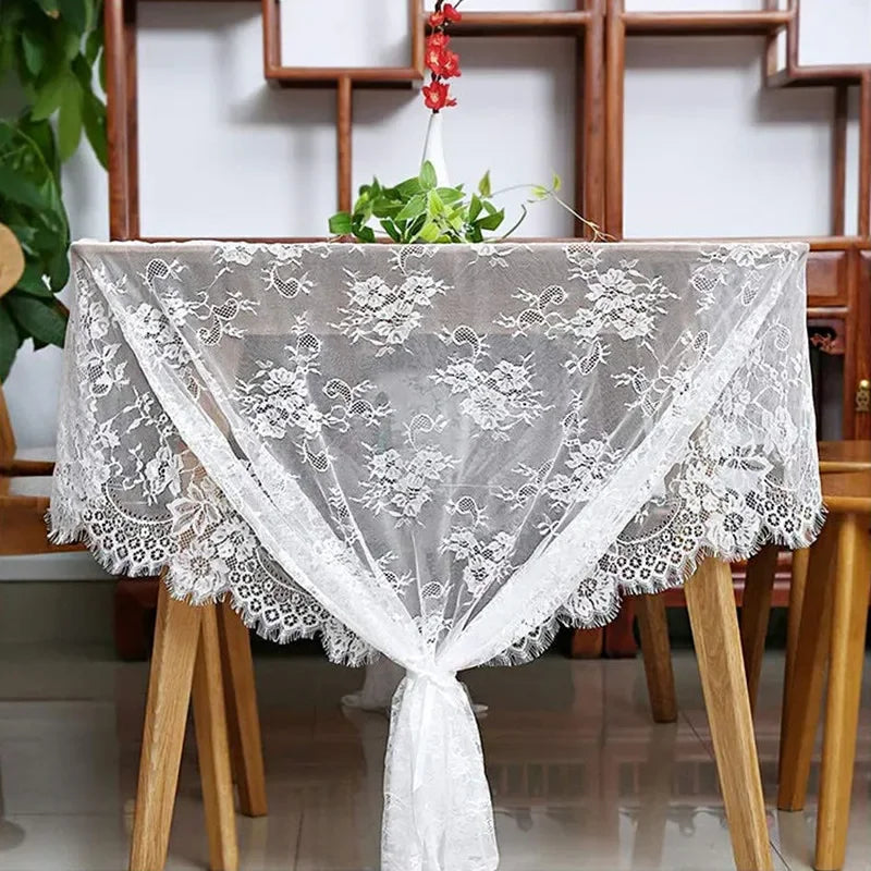 A round wooden table adorned with a sheer white lace tablecloth featuring floral patterns. A small potted plant with a red flower is placed on top, part of a decorative shelf backdrop.