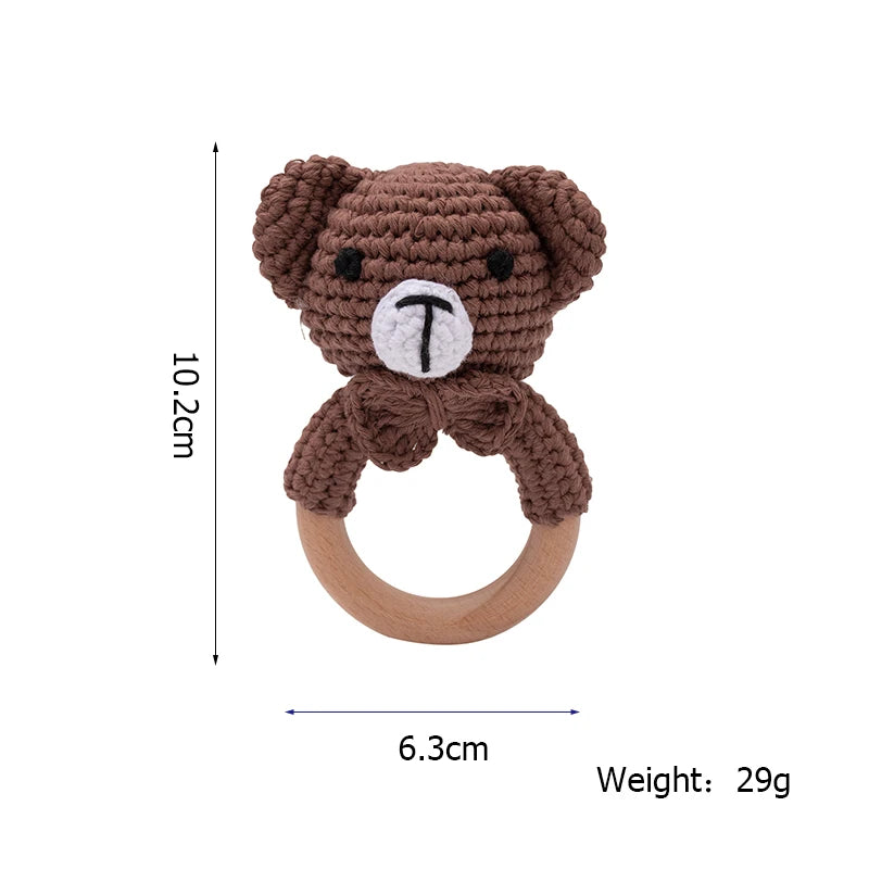 Brown crocheted teddy bear rattle with a wooden ring. The bear's head and arms are attached to the top. Dimensions: 10.2 cm tall, 6.3 cm wide, weighing 29 grams.