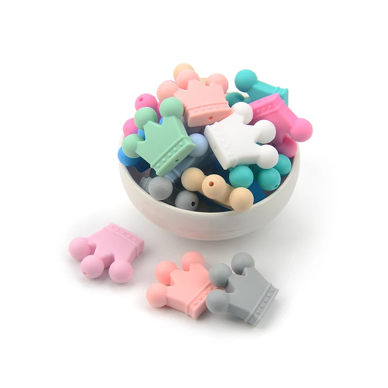 A white bowl filled with colorful silicone beads shaped like crowns, in shades of pink, green, white, gray, and blue. Additional beads are scattered around the bowl.