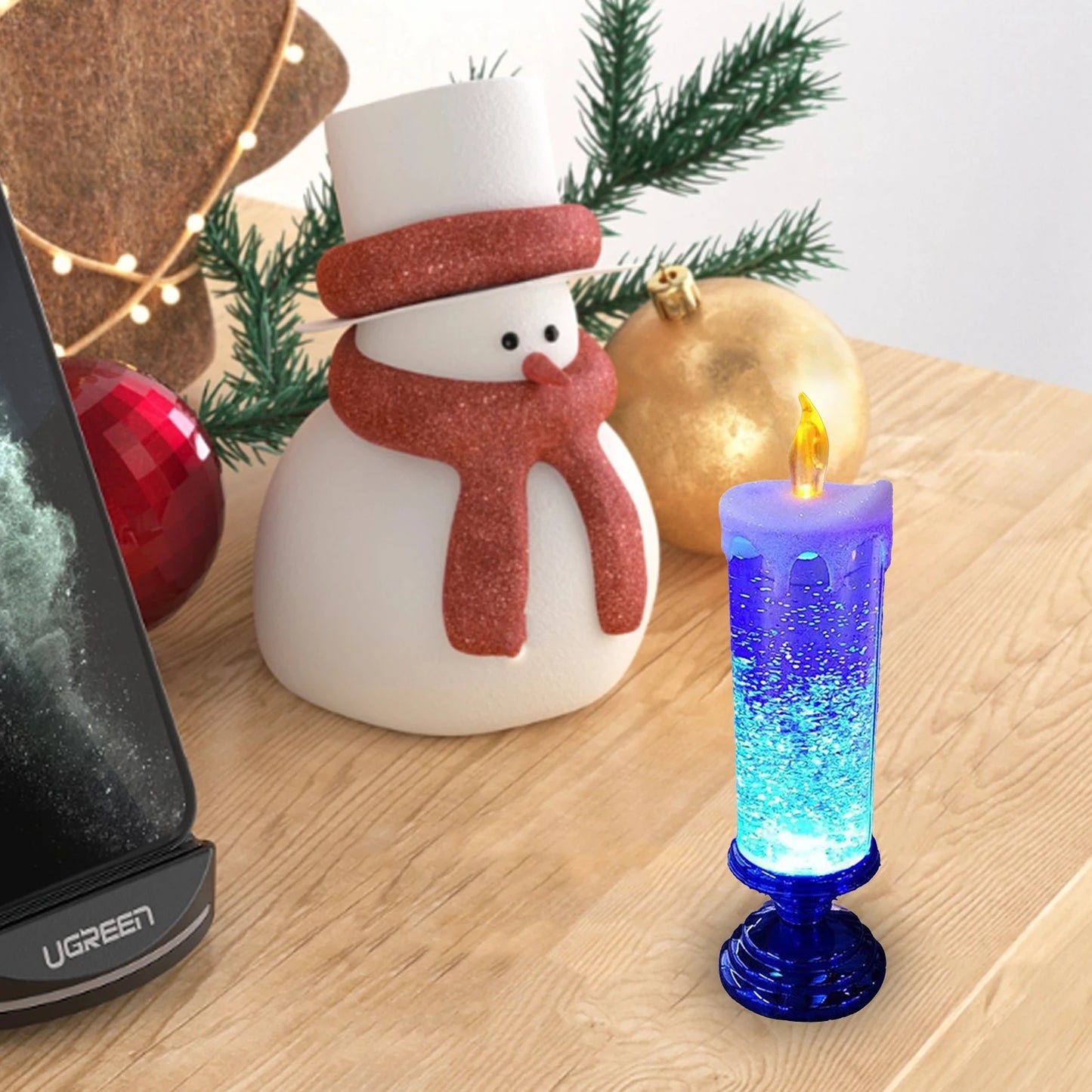 A snowman figurine with a red scarf stands beside festive decorations, including a shimmering blue LED candle and Christmas ornaments on a wooden surface.