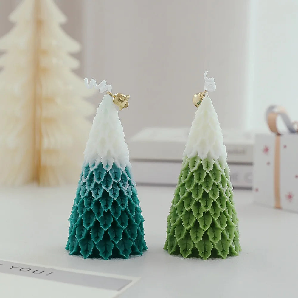 Two Christmas tree candles with layered textures, one in green and the other in blue, topped with small gold bells. Blurred background with soft holiday decor elements.