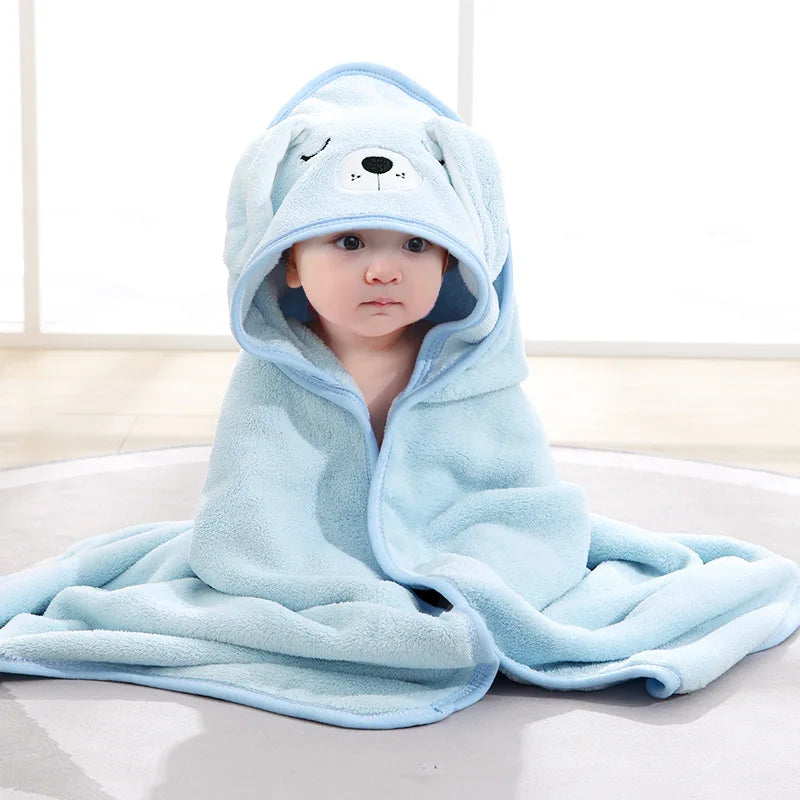 A baby sits on a gray mat wearing a fluffy blue hooded towel with bear ears and facial features. Bright room with natural light streaming in from a large window.