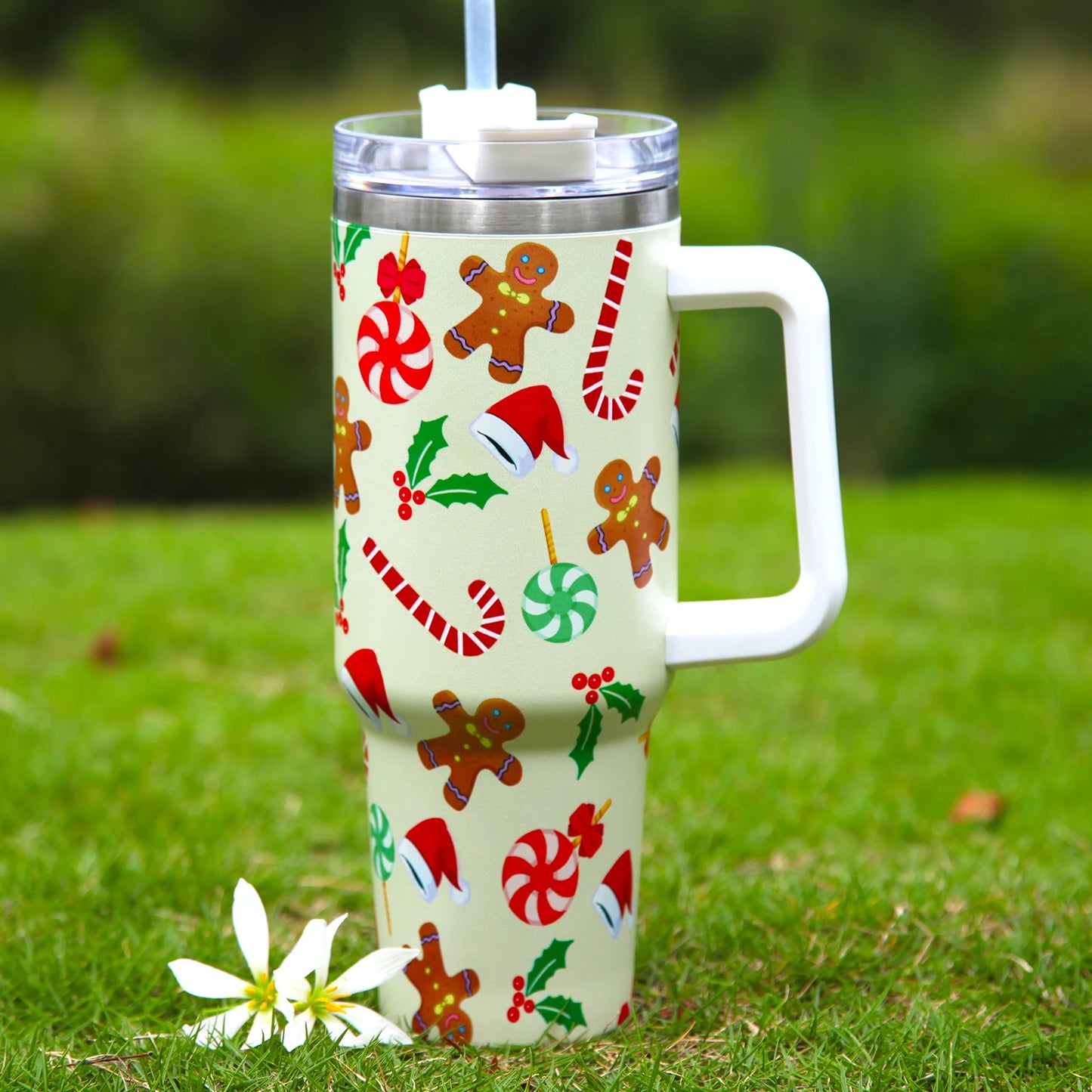 Festive travel mug with gingerbread men, candy canes, holly, and Santa hats on a grassy background. A white flower lies nearby.