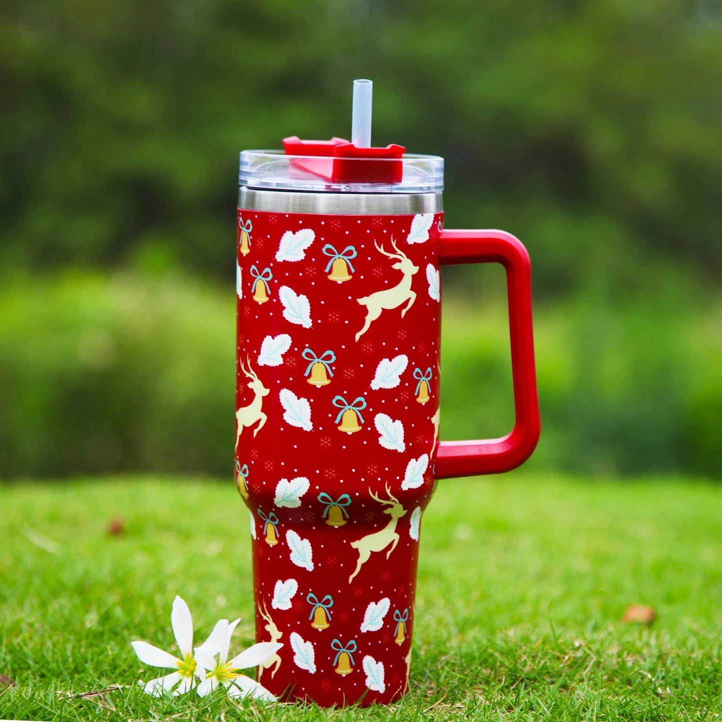 Red tumbler with reindeer, bells, and leaf designs, featuring a handle and straw. It sits on grass with white flowers nearby, against a blurred green background.