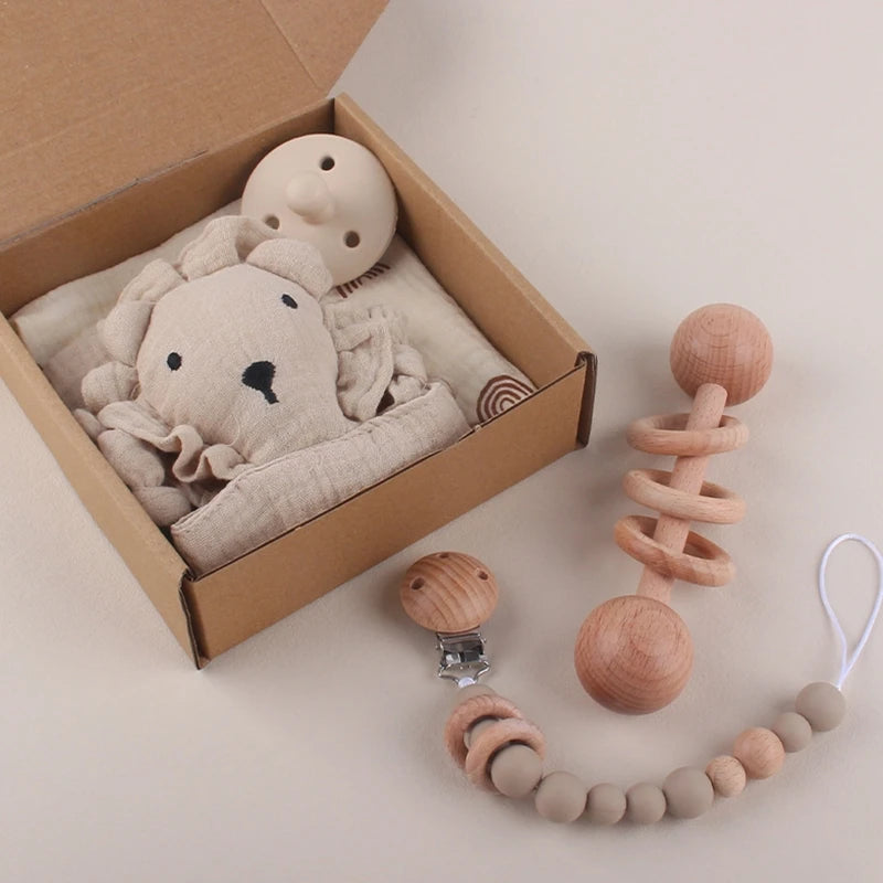 A cardboard box contains a beige stuffed lion toy and a small beige blanket. Next to it, a wooden baby rattle and a pacifier clip with wooden and silicone beads are displayed.