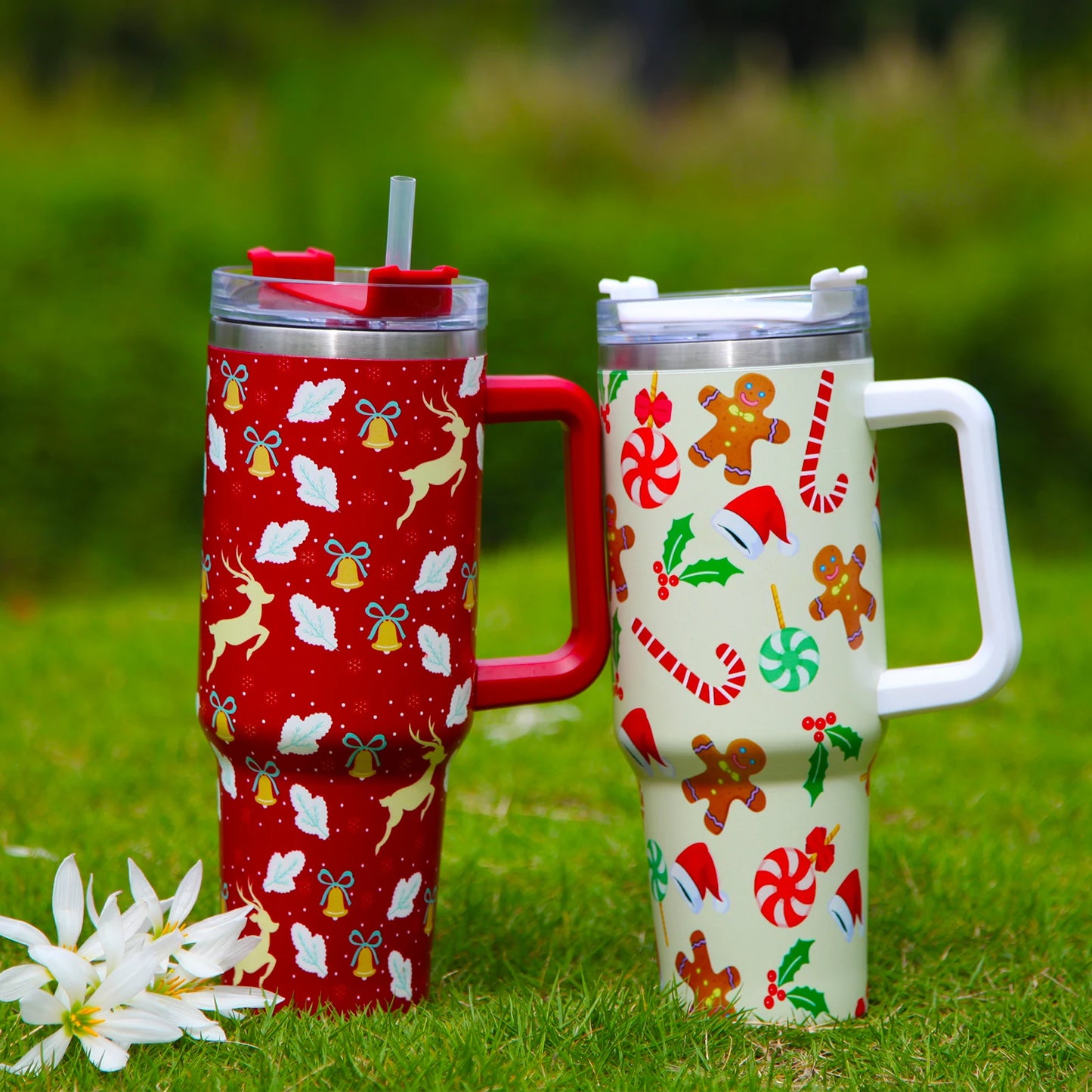 Two festive travel mugs with lids and handles on grass. The red mug features reindeer and bells, while the cream mug displays gingerbread men and candy canes. White flowers are beside them.