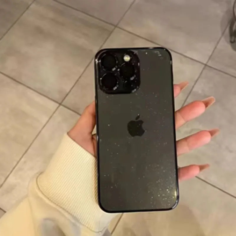 A person holding a dark-colored iPhone with a sparkling case, showing the triple camera setup. The phone is held above a tiled floor, with the person's sleeve visible.