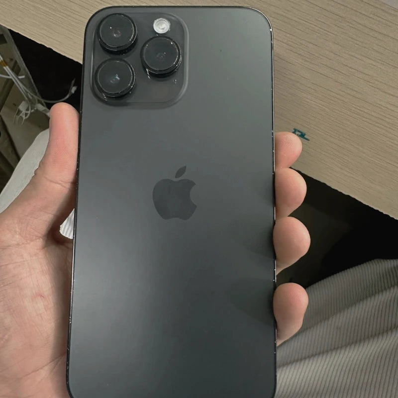 A person holds a dark-colored smartphone with three large camera lenses on the back. It's resting in a hand, and the Apple logo is visible. The phone is against a wooden surface.
