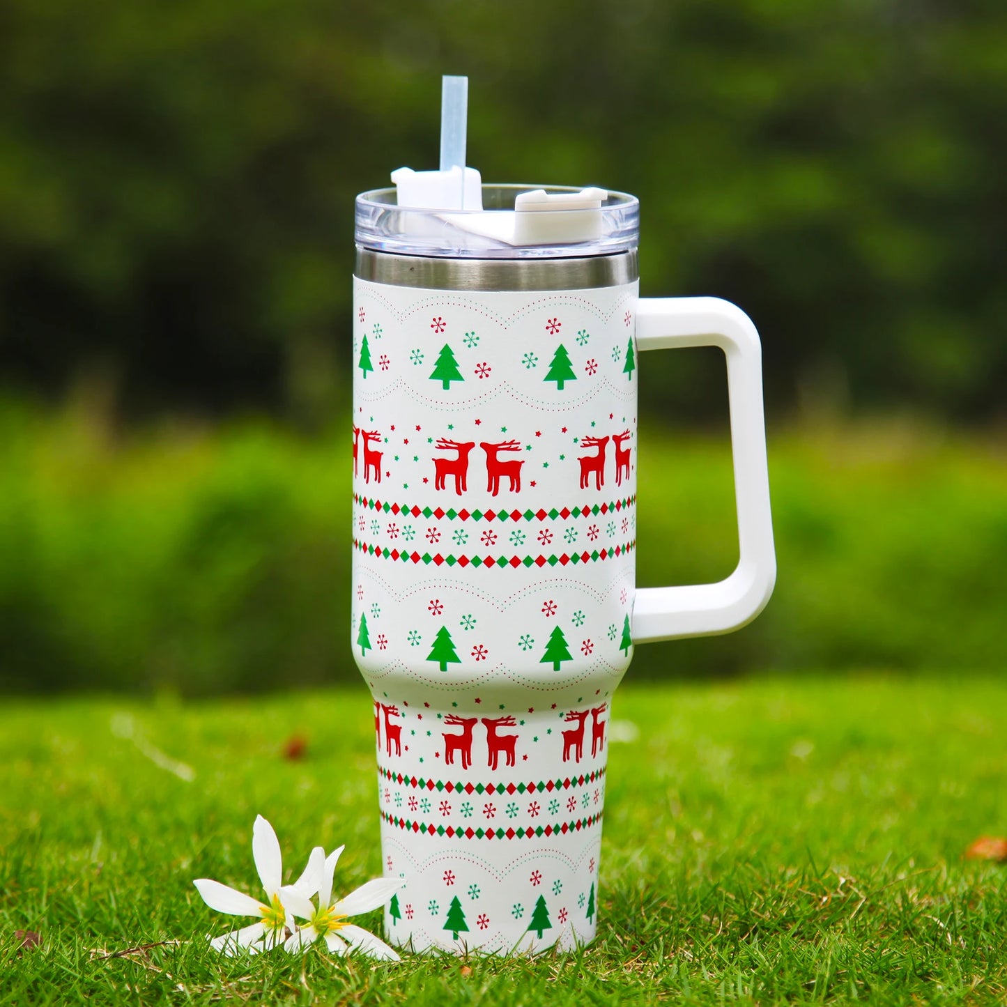 Festive travel mug with reindeer and tree patterns, featuring a lid and straw, sits on green grass next to a white flower. The background is a blurred greenery.