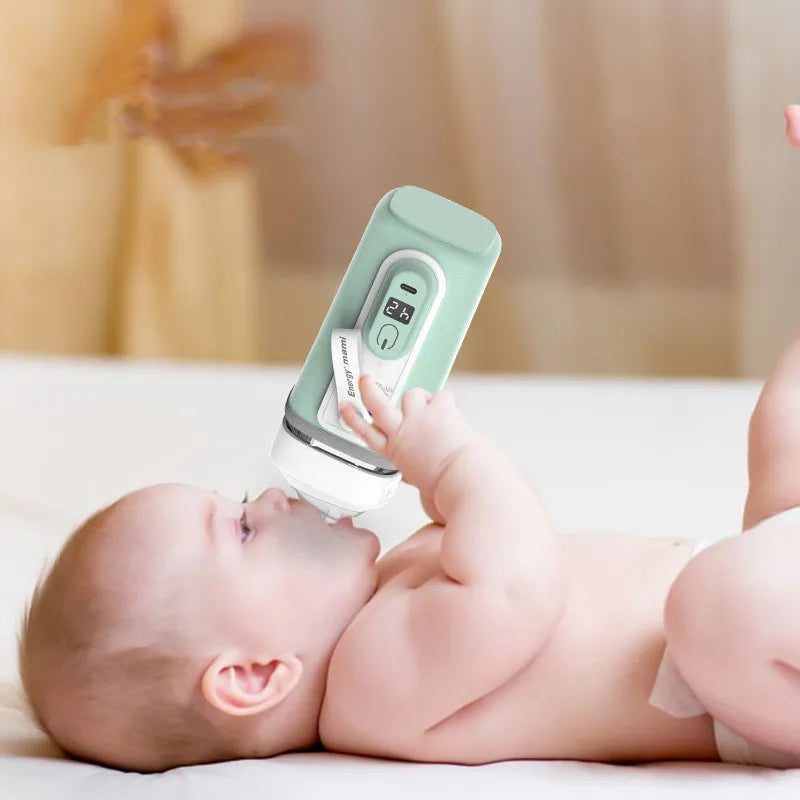 A baby lies on a bed, holding a modern baby bottle with a digital display showing "24" in one hand, drinking from the bottle. The bottle has a light green cover.