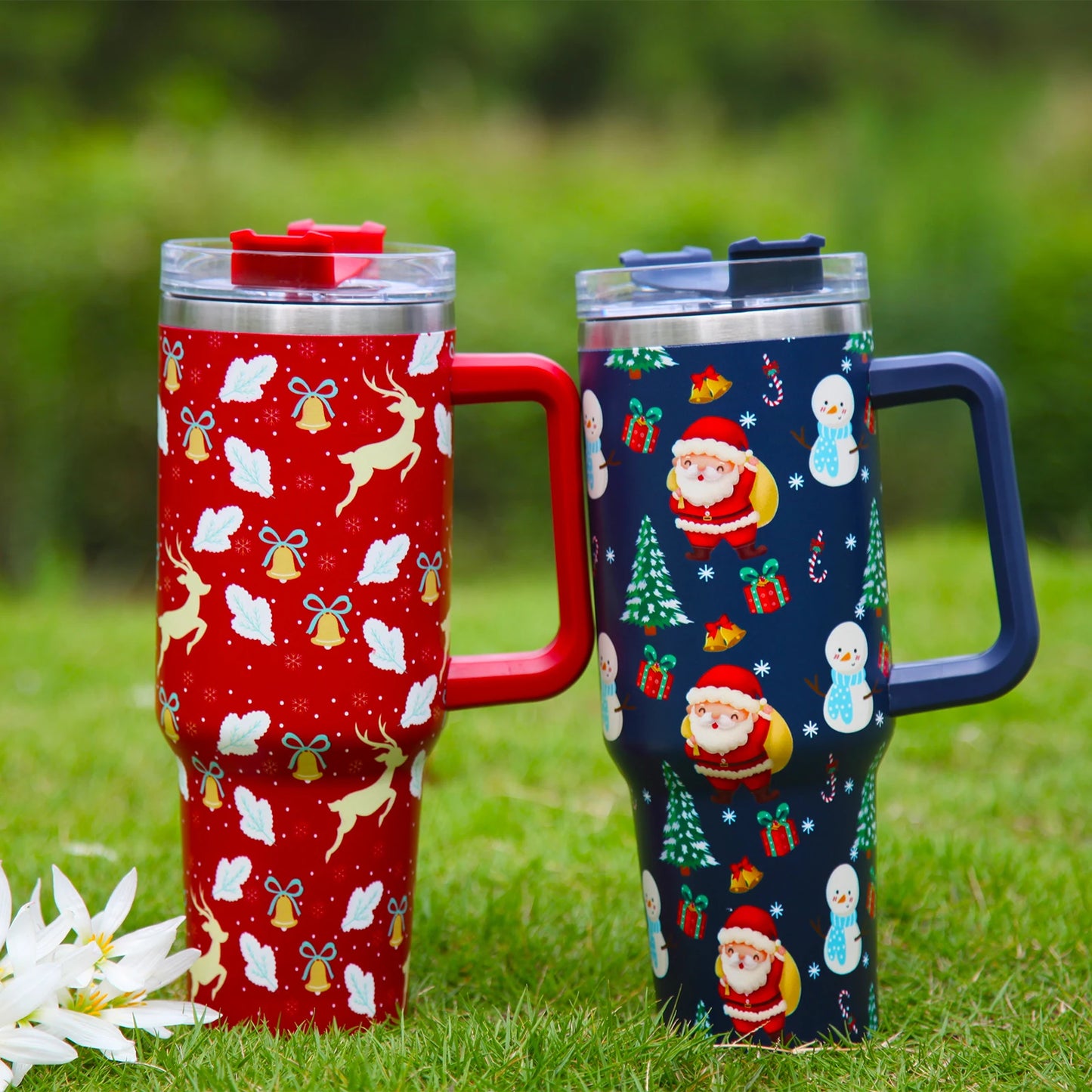 Two festive travel mugs on grass: one red with reindeer and bells, the other blue with Santa, snowmen, and trees. Both have clear lids and matching handles.