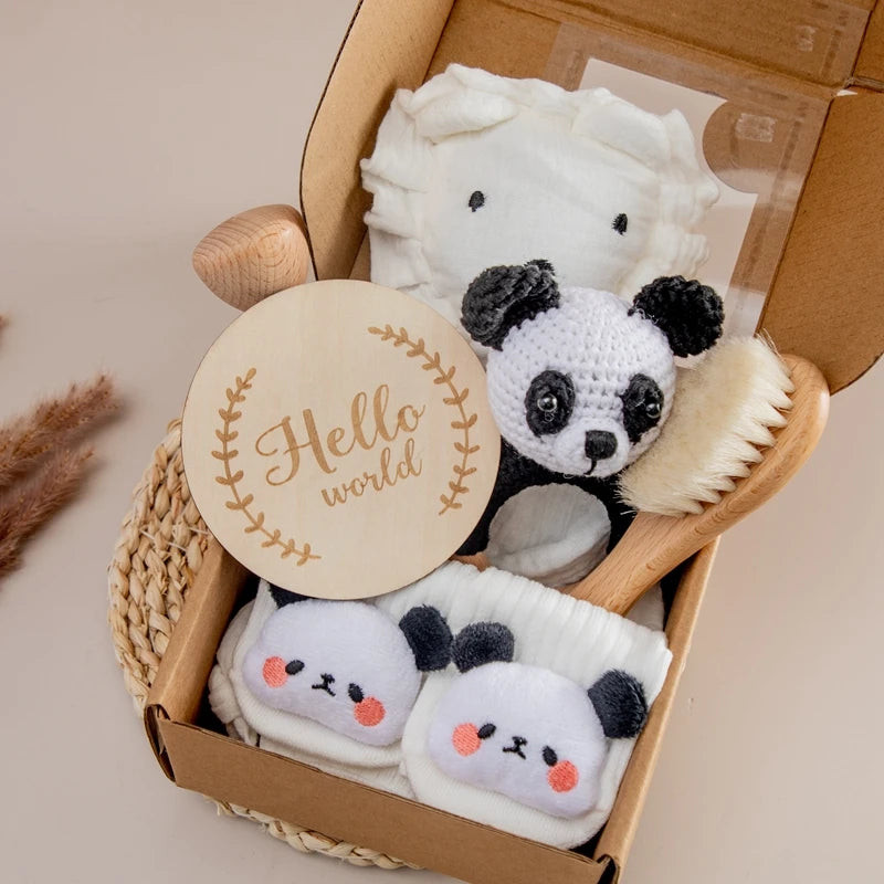A gift box containing a crocheted panda toy, a wooden hairbrush, cozy socks with panda faces, a soft white blanket, and a "Hello world" wooden sign, placed on a woven mat.