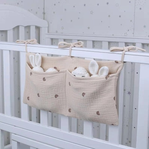 Hanging fabric storage on a white crib with two pockets holding plush bunny toys. The organizer is beige with small brown patterns and is tied with bows for attachment.