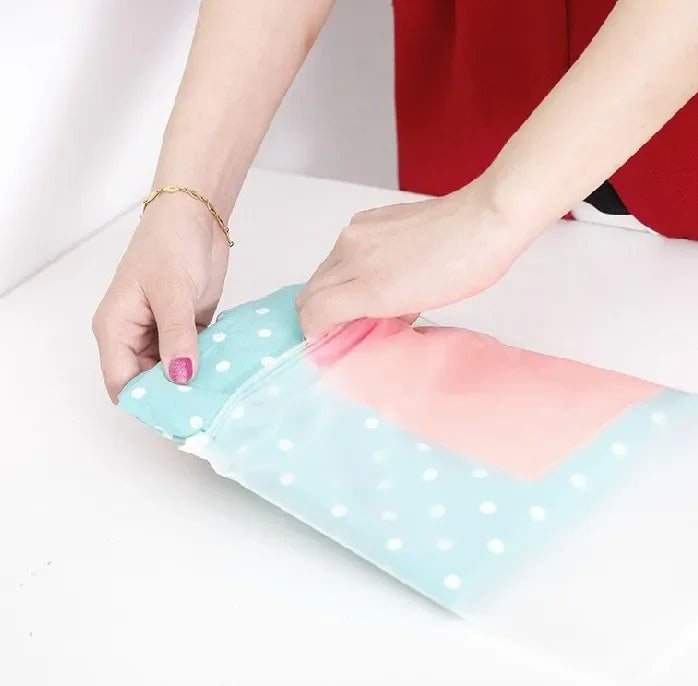Hands folding a light blue polka dot fabric bag with a pink section on a white surface. The person wears a red sleeve and gold bracelet.