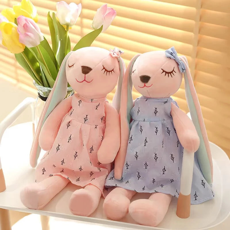 Two plush bunny dolls with closed eyes, wearing pink and blue dresses with black patterns, sit on a white surface. A vase with tulips is in the background.