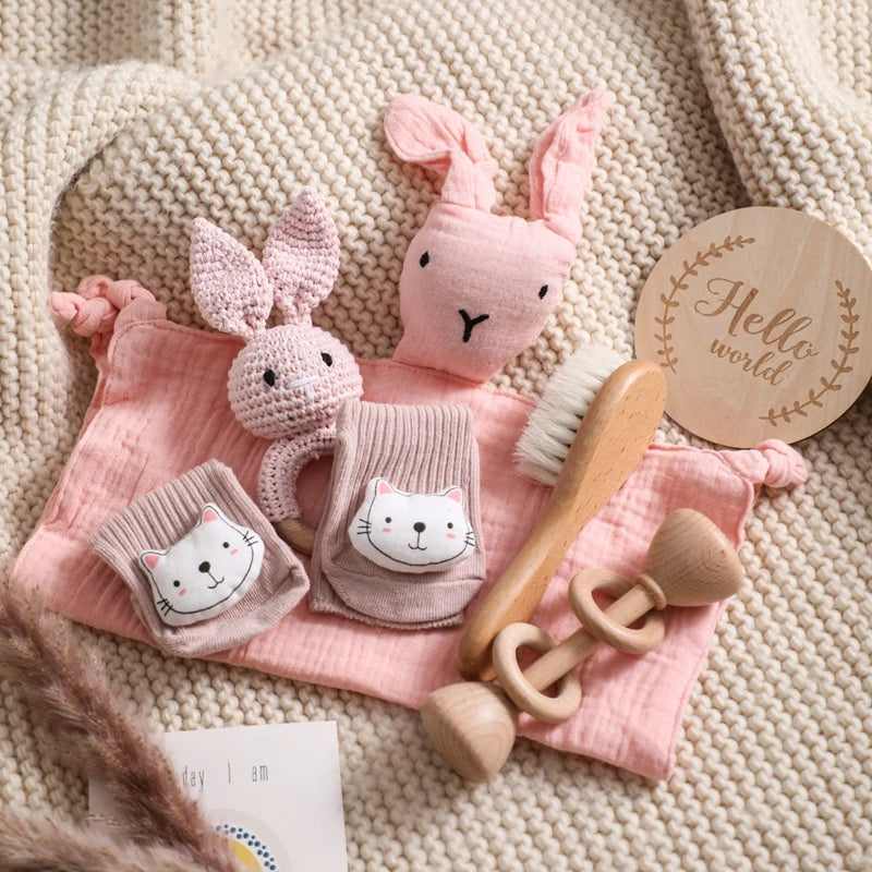 Baby items on a knitted blanket: pink bunny toys, knitted socks with cat faces, a wooden hairbrush, and wooden rattles. A wooden plaque reads "Hello world."