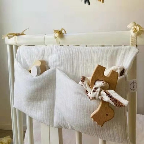 A white crib organizer with two fabric pockets hangs on a crib rail. One pocket holds wooden toys, and the other contains a stuffed toy. Beige ribbons secure it to the crib.