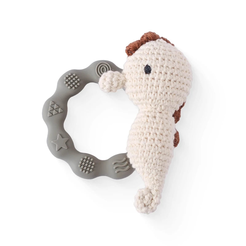 A crocheted seahorse toy with a brown mane is attached to a textured gray silicone teething ring. The ring features various embossed shapes like stars and circles.