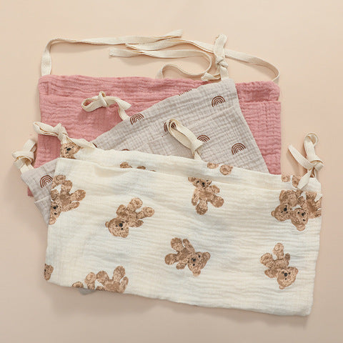 Three baby bibs with ties are layered on a beige background. The top bib is cream with teddy bear prints, the middle is grey with rainbows, and the bottom is pink.