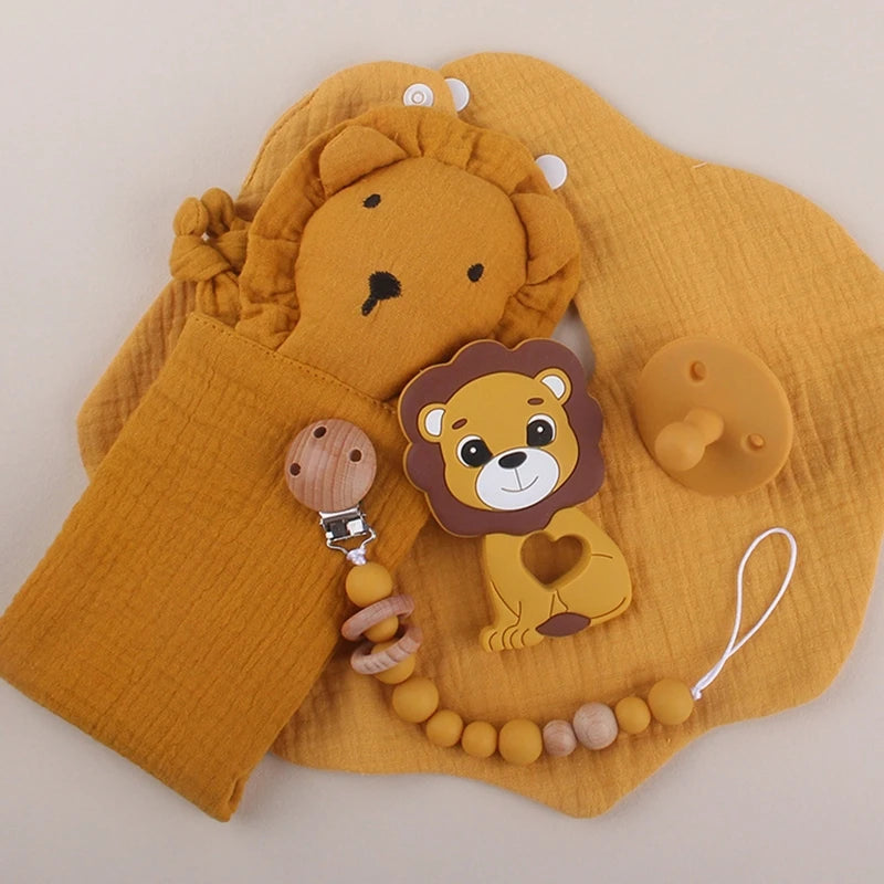 A collection of lion-themed baby items: a plush lion mitt, lion teether with heart cutout, pacifier, and a clip featuring wooden and yellow beads, all on a matching mustard cloth.