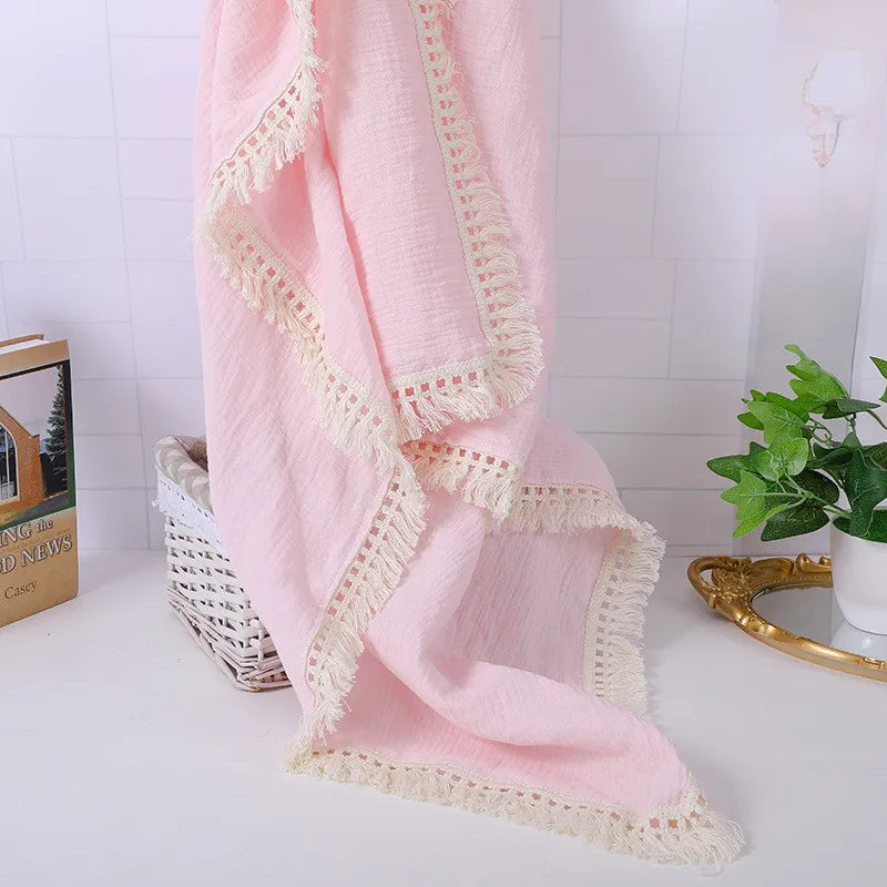 Soft pink fabric with intricate lace edges drapes over a white woven basket. Beside it are a book and a small potted plant, creating a cozy, elegant display on a white surface.