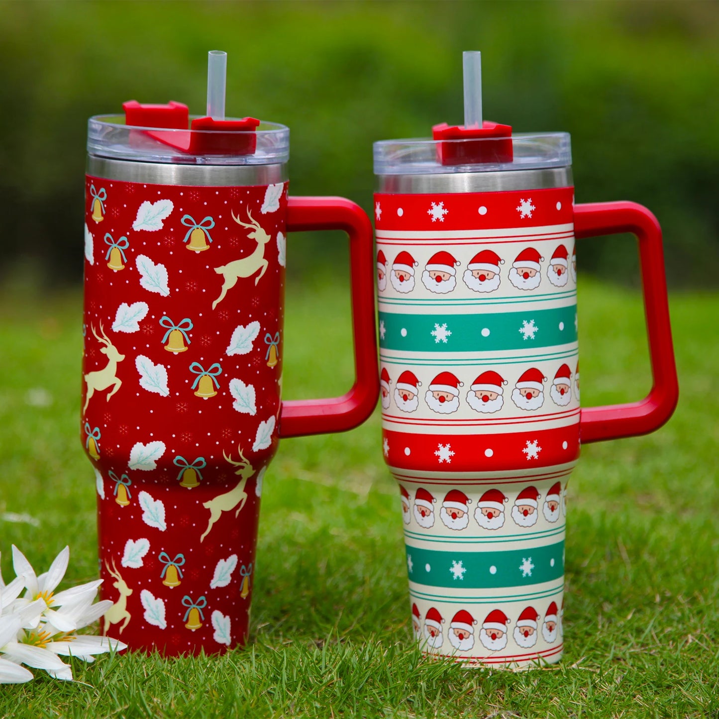 Two festive mugs with straws on grass. One red with reindeer and bells, the other with Santa and snowflakes in red and green. White flowers nearby.