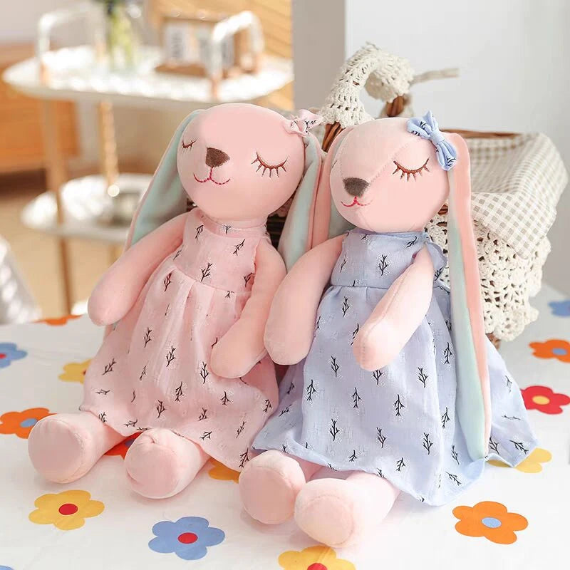 Two plush toy rabbits in pastel dresses, one pink and one blue, sit on a floral-patterned tablecloth. Both have closed eyes and long ears, with bows on their heads.