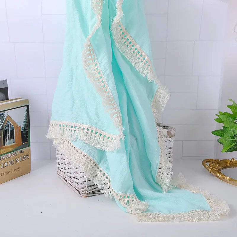 A light blue woven blanket with cream fringed edges draped over a white wicker basket. A book and green plant sit on a white tiled surface in the background.