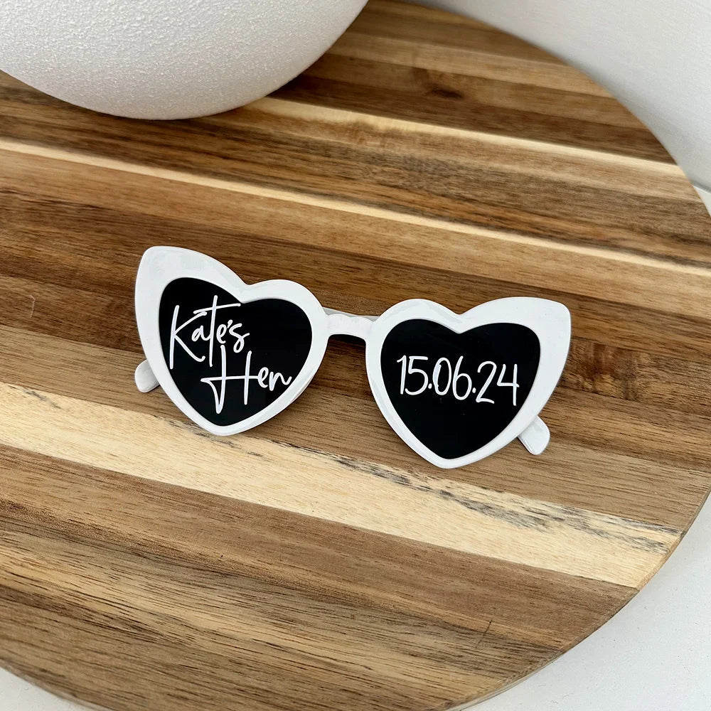 Heart-shaped sunglasses with white frames on a wooden surface. Left lens reads "Kates Hen" and right lens shows the date "15.06.24".