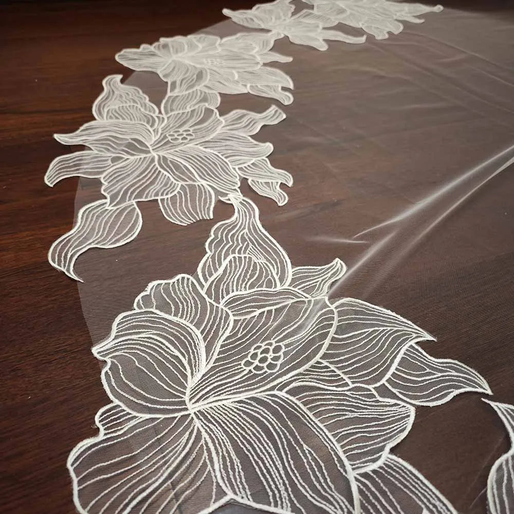 Delicate white lace fabric with intricate floral patterns laid on a wooden surface, showcasing detailed petals and leaves in an elegant design.