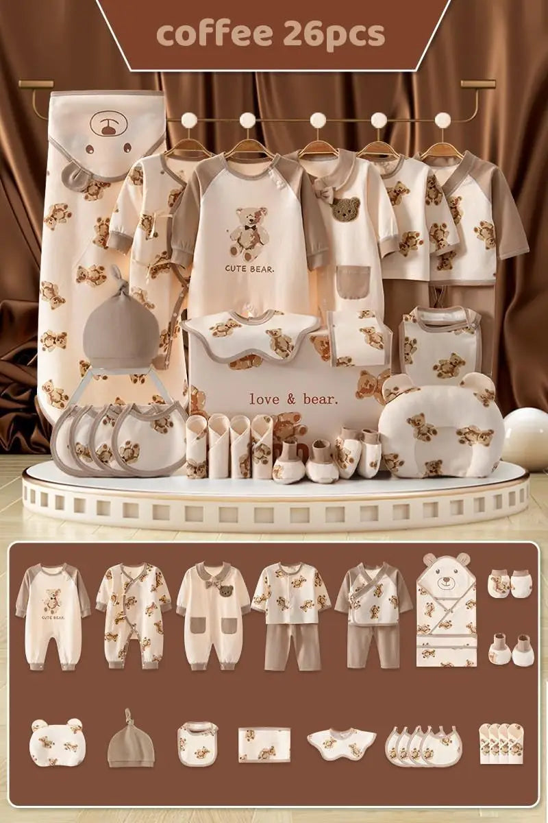 A 26-piece baby clothing and accessory set in a coffee color theme with teddy bear designs. Includes onesies, bibs, hats, a blanket, and towels, displayed on a circular platform.