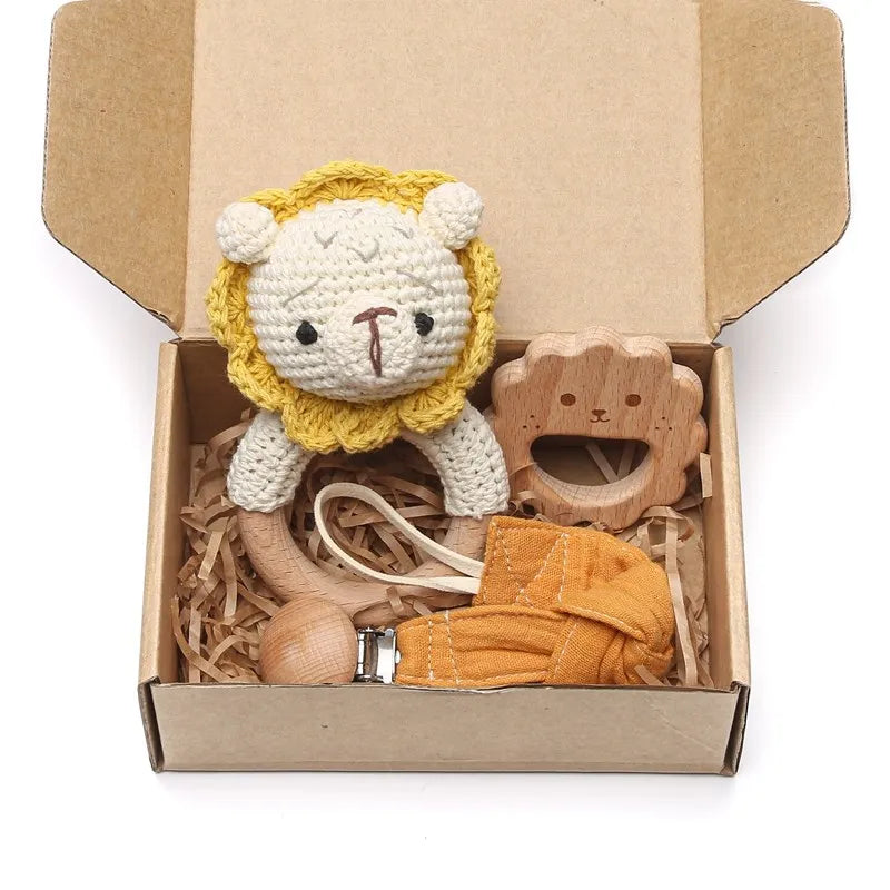 Open cardboard box containing a knitted lion toy, a wooden teething ring, and an orange fabric pacifier clip set on shredded paper packing material.