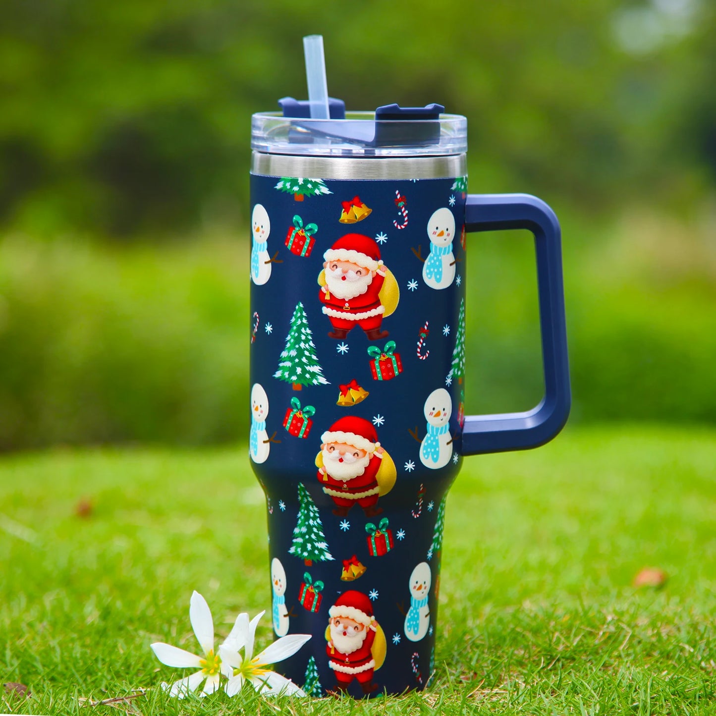 Festive tumbler with Santa, snowmen, and gift patterns on a blue background, set on grass with a white flower. Includes a handle and clear lid with straw.
