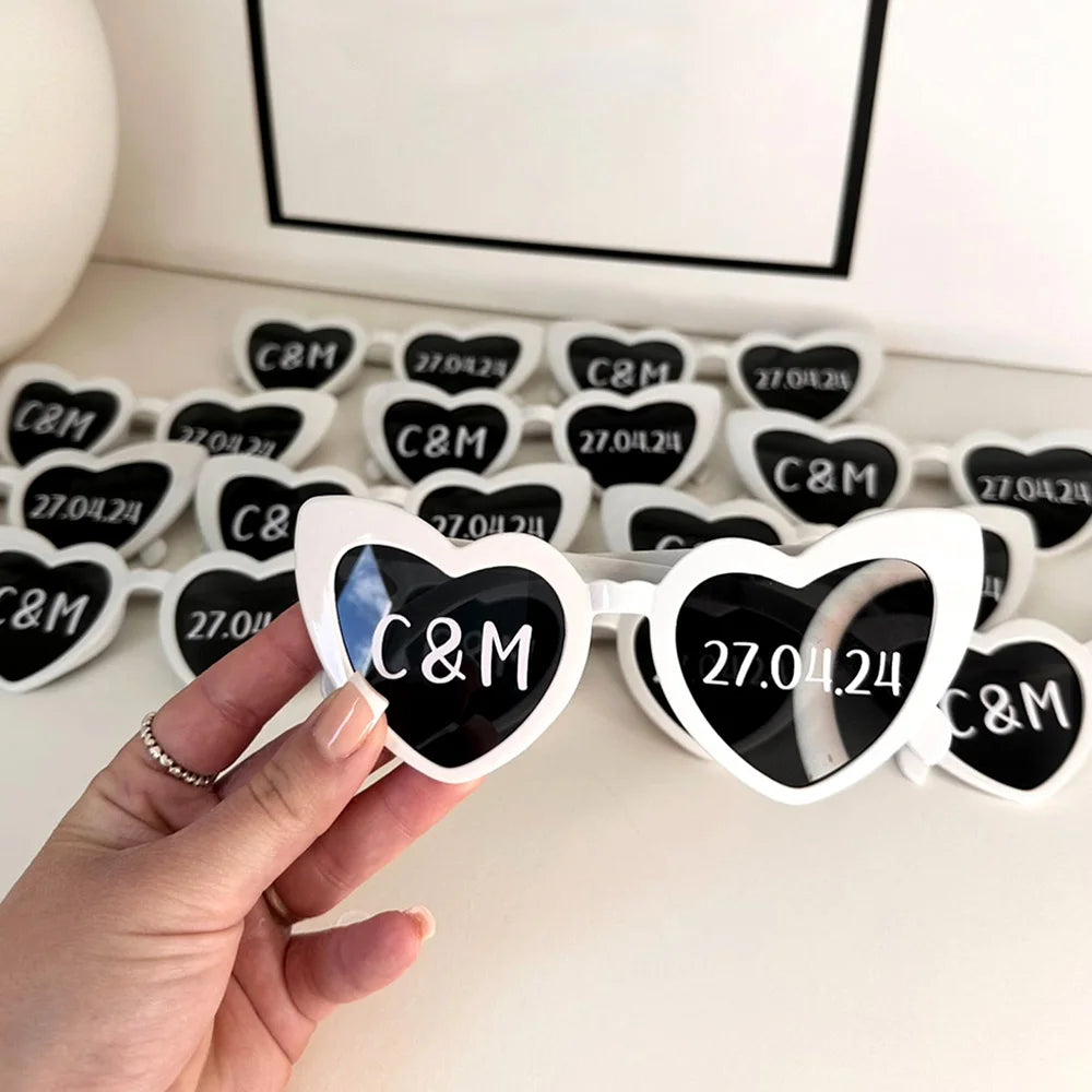 A hand holds white heart-shaped sunglasses with "C&M" on one lens and "27.04.24" on the other. Several identical sunglasses are displayed in the background.