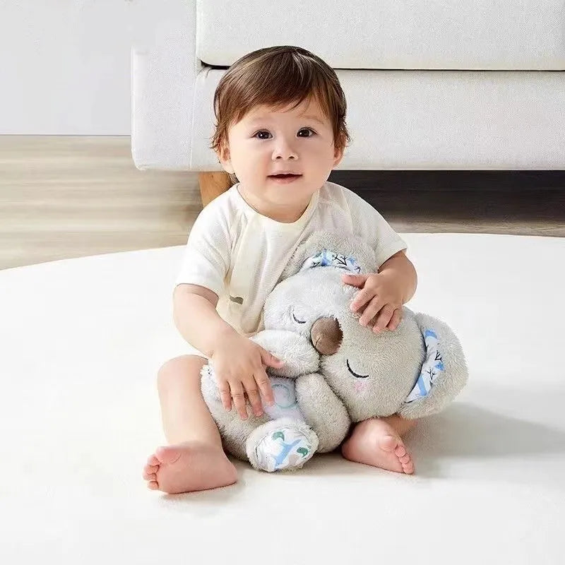 A toddler sits on a white rug holding a large, plush koala bear. The child wears a light-colored outfit and has short brown hair. A gray sofa is in the background.