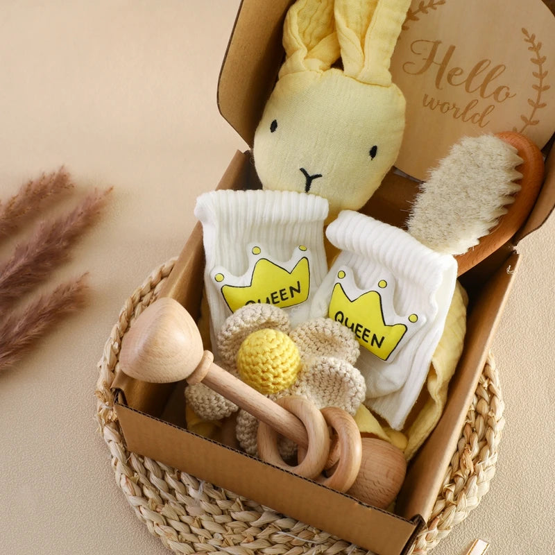 A gift box with a yellow bunny plush, baby socks with "Queen" and crown prints, a wooden rattle, a knitted flower, and a soft brush. A "Hello world" card is included.