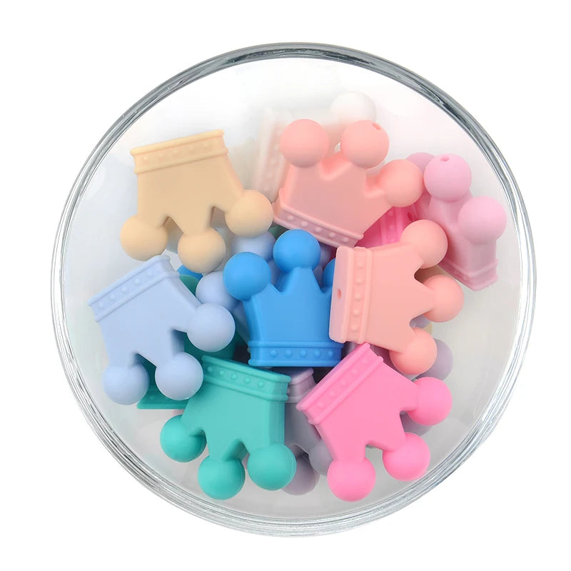 A glass bowl filled with colorful, pastel crown-shaped silicone teething toys in shades of pink, blue, green, and beige.