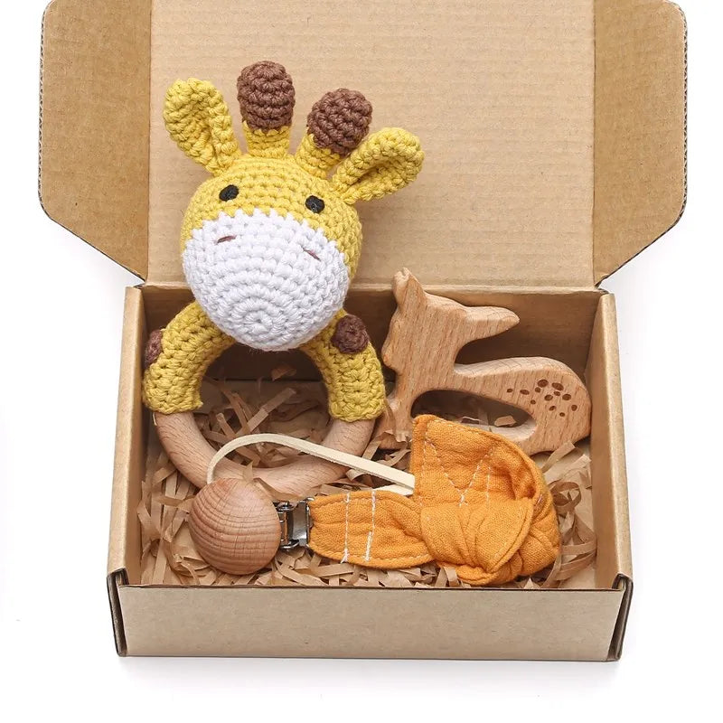 A gift box with a crocheted giraffe rattle, wooden deer toy, pacifier clip, and a yellow fabric leaf. Set on shredded brown paper inside a cardboard box.