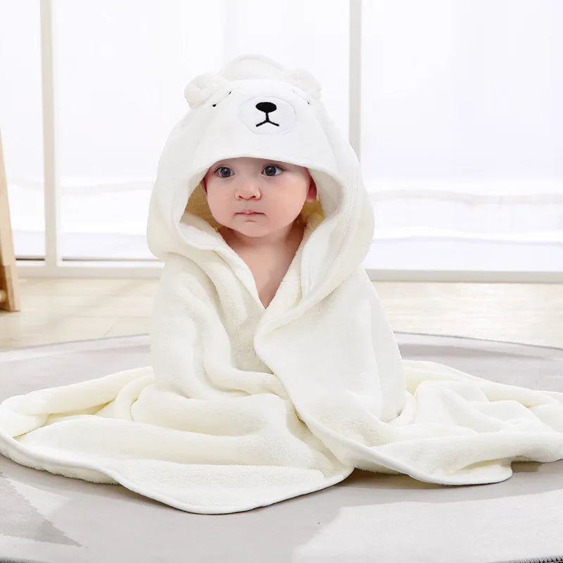 A baby wrapped in a white fleece blanket with a bear hood, sitting on a light gray circular rug. The hood features a bear face design with ears, nose, and eyes.