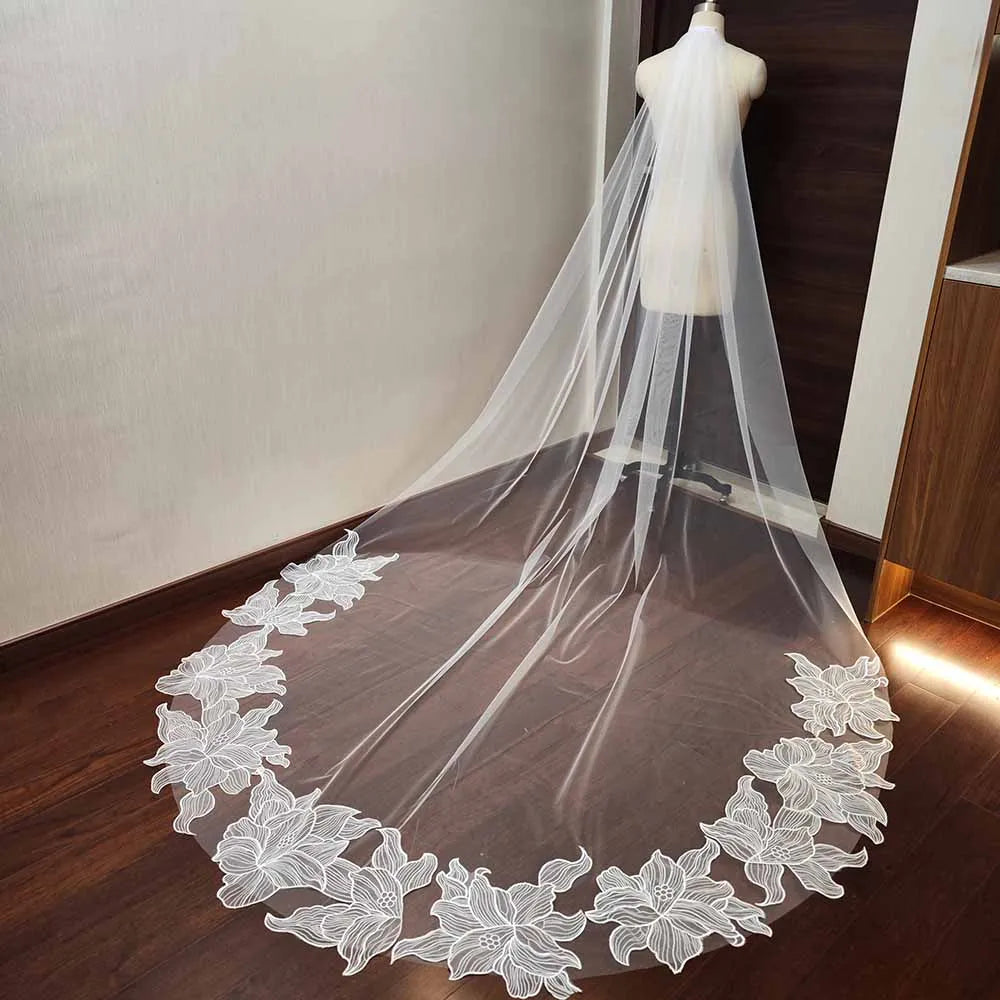 A long, sheer bridal veil with intricate white floral lace edging displayed on a mannequin, flowing elegantly across a wooden floor.