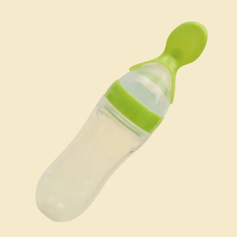 Transparent baby feeding bottle with a green silicone spoon attachment, designed for easy feeding. The bottle has a green cap and is set against a plain beige background.