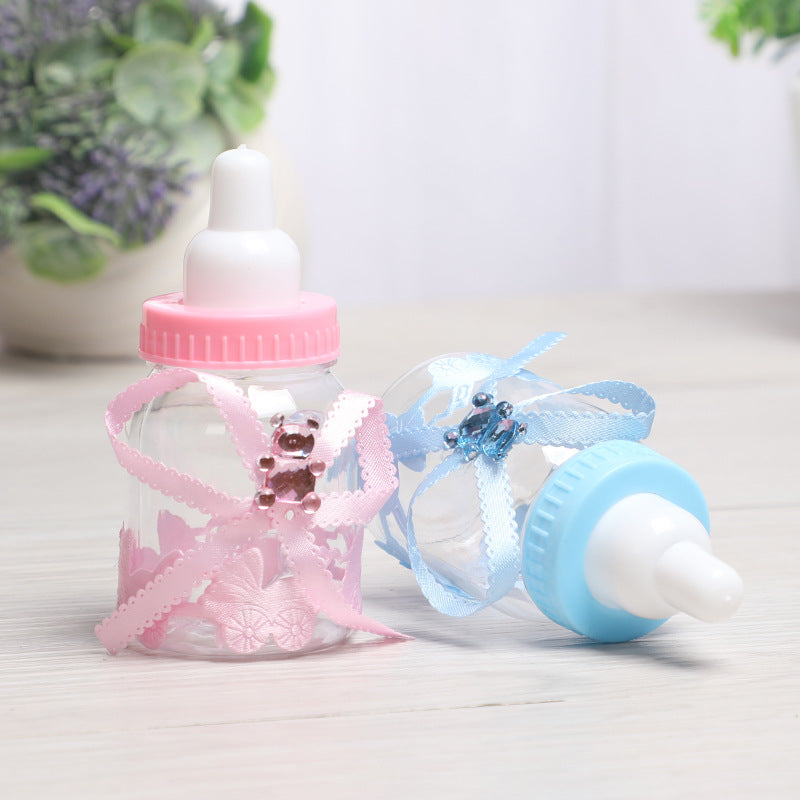 Two decorative baby bottles with white nipples; one has a pink cap and ribbon with a teddy bear embellishment, the other has a blue cap and ribbon, placed on a light wooden surface.