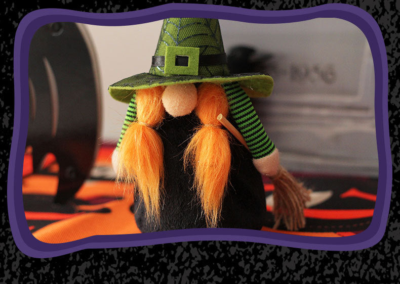 A small stuffed witch figurine with a green hat and long orange braids stands on a Halloween-themed table. The witch's outfit is striped green and black, and she holds a tiny broomstick. The scene is framed with a purple border, and a blurred black object is visible in the background.