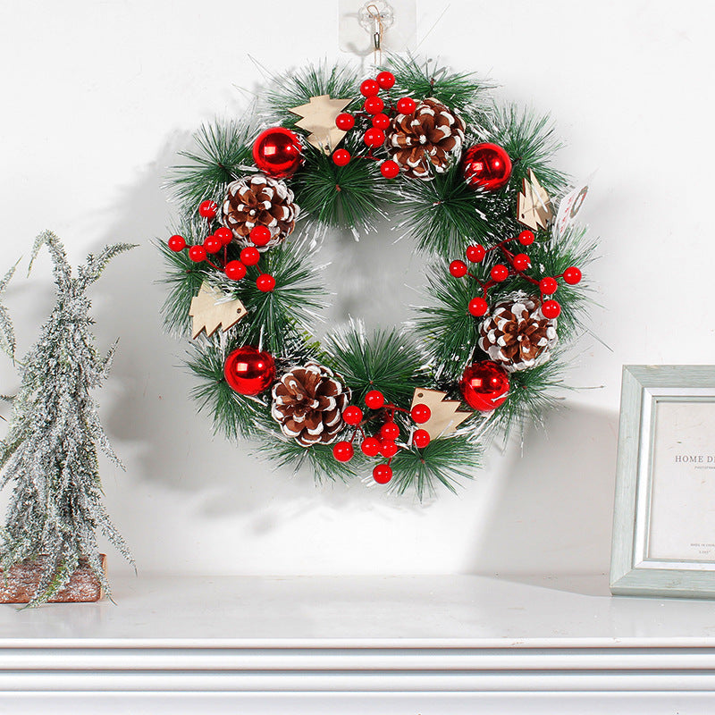 Festive wreath on a white wall, featuring green foliage, red berries, pine cones, and red ornaments. A snow-dusted tree decoration sits on a nearby shelf with a framed picture.
