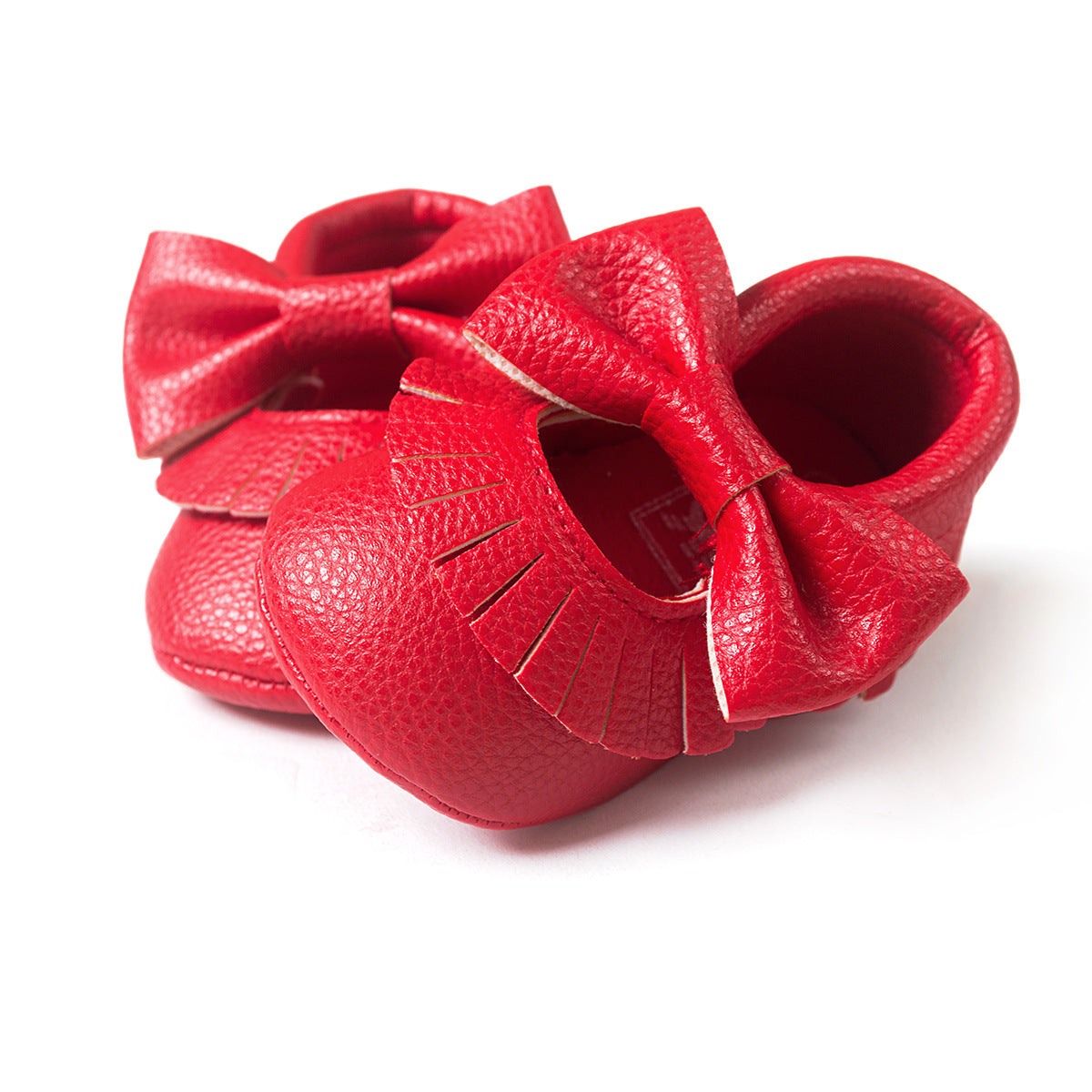 Red baby shoes with textured leather and oversized bows on the front. The shoes feature small fringe detailing, creating an adorable and elegant look, set against a white background.