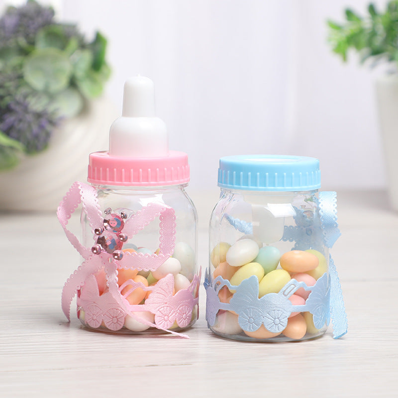 Two baby-themed jars filled with colorful candies. One has a pink lid and bow, while the other has a blue lid and bow. Decorative lace features baby carriage designs.