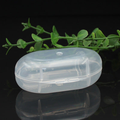 A transparent, oval-shaped soap case rests on a glossy black surface with green leaves in the background. The case has visible hinges and a snap closure, designed to securely hold soap.