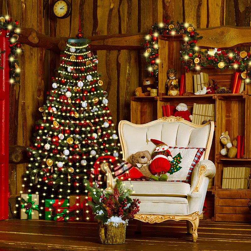 Cozy room with decorated Christmas tree, luminescent garlands, and gifts. A plush chair holds festive pillows and toys. Shelves with books and ornaments enhance the warm, rustic atmosphere.