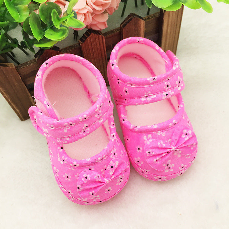 Pink baby shoes with floral patterns and bows on the toes, placed on a light surface. A wooden planter with green leaves and pink flowers is in the background.