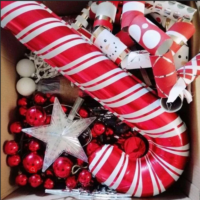 A box filled with Christmas decorations, including a large candy cane with red and white stripes, red baubles, white orbs, a silver star, and various paper tubes.